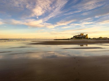 Bamburgh Castle as seen across Bamburgh Castle Beach. It looks like dusk, with a pale golden sky on the horizon and the colours reflecting off the water on the sand.