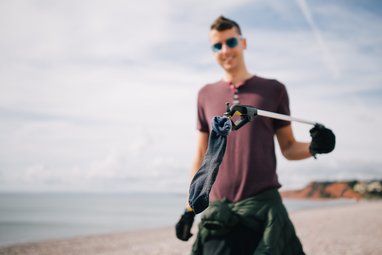 A man holding up a dark coloured sock up to the camera. He is using a litter picker to hold it. The sock is dark in colour and covered with sand.