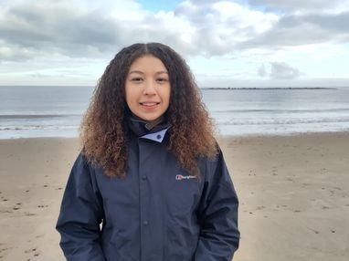 Trustee Rebecca smiles on a sandy beach with the ocean behind her. It looks like an overcast UK day. She has curly brown hair and is wearing a navy waterproof jacket.