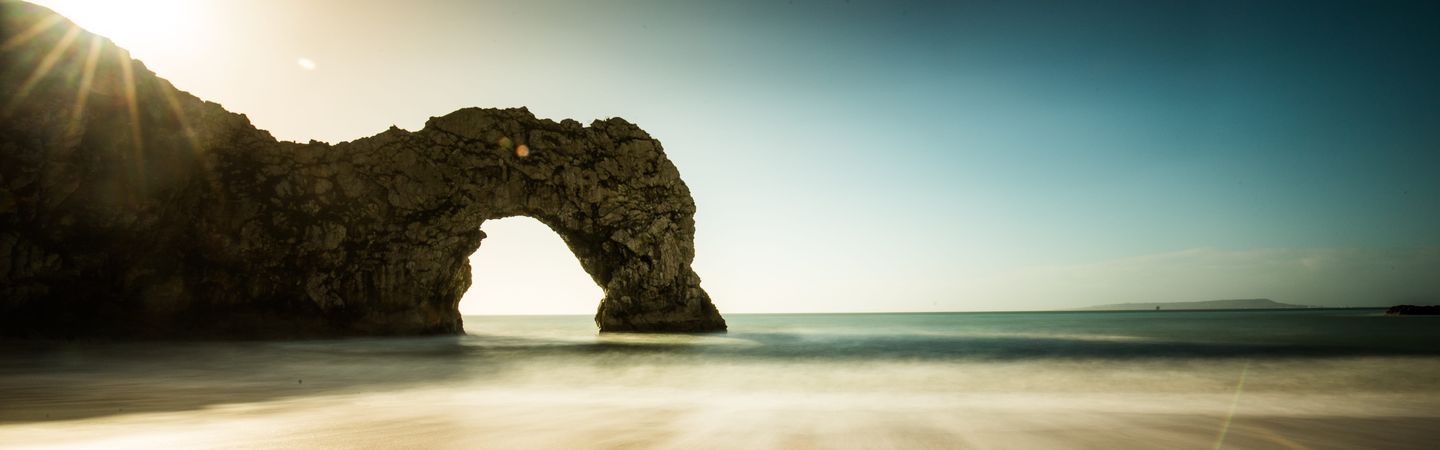 A long exposure photograph of the natural limestone arch known as Durdle Door on the Jurassic Coast in Dorset, England, with the sun low in the sky and blurred waves on the sand.
