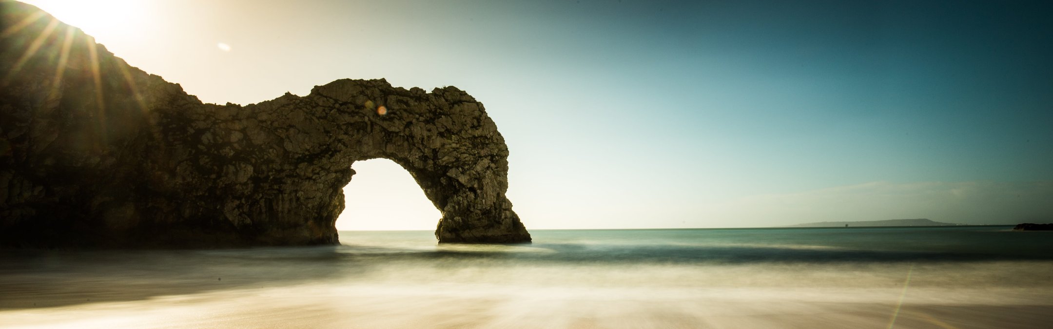 A long exposure photograph of the natural limestone arch known as Durdle Door on the Jurassic Coast in Dorset, England, with the sun low in the sky and blurred waves on the sand.