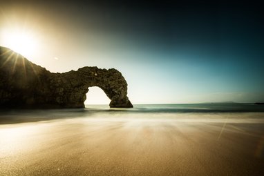 A long exposure photograph of the natural limestone arch known as Durdle Door on the Jurassic Coast in Dorset, England, with the sun low in the sky and blurred waves on the sand.