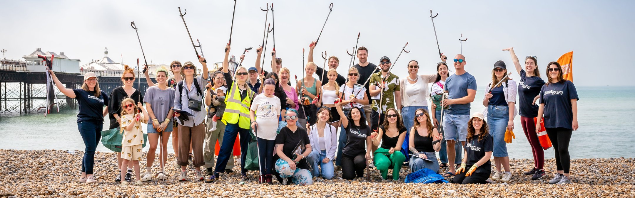 A large group of approximately 30 volunteers participating in a beach clean event stand smiling on a pebble beach, many holding litter pickers and bags, with a pier visible in the background.