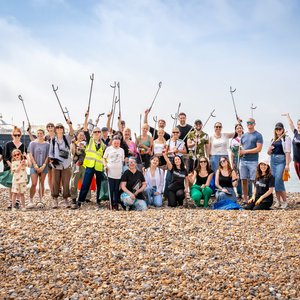 A large group of approximately 30 volunteers participating in a beach clean event stand smiling on a pebble beach, many holding litter pickers and bags, with a pier visible in the background.