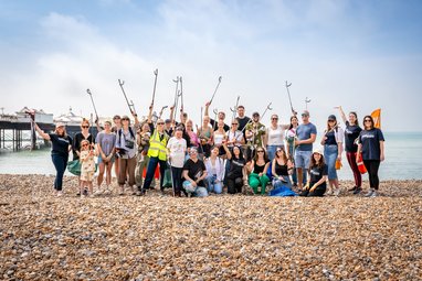 A large group of approximately 30 volunteers participating in a beach clean event stand smiling on a pebble beach, many holding litter pickers and bags, with a pier visible in the background.