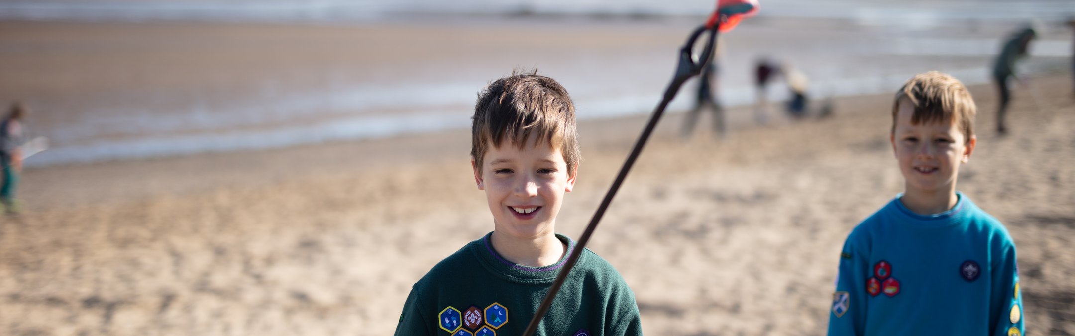 2 boys dressed in the Scouts uniform standing on a beach. One boy is holding up marine litter with a litter picker.