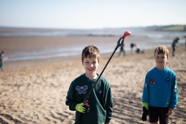 2 boys dressed in the Scouts uniform standing on a beach. One boy is holding up marine litter with a litter picker.