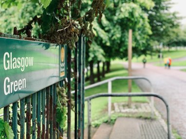 A close-up of a green metal sign that reads "Glasgow Green" in white lettering, attached to a weathered green iron fence. Behind the sign, a paved pathway leads into a lush, tree-lined park where blurred figures can be seen walking in the distance.