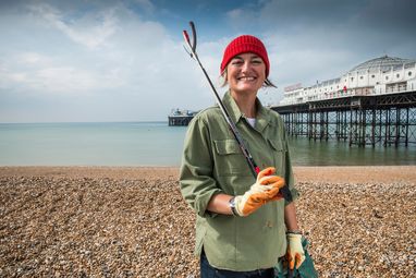 Zoe Lyons stands smiling on Brighton beach with the pier in the background. She is holding a litter grab over her shoulder and is wearing a khaki shirt and red beanie hat.
