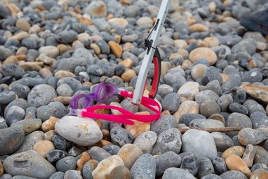 A pair of pink swimming goggles being held up by a litter picker on a pebble beach