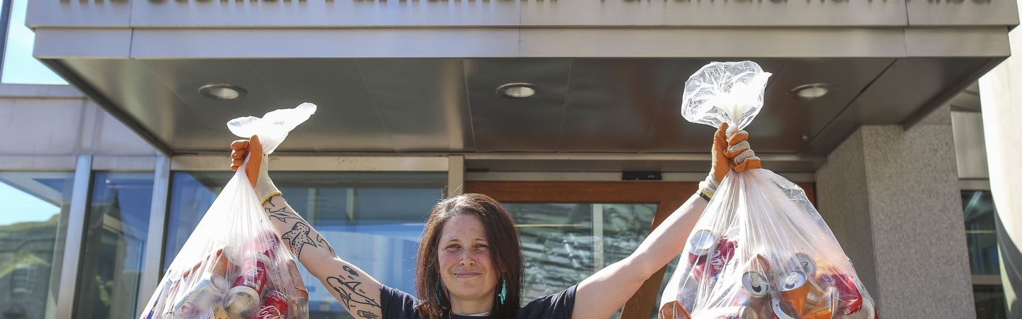 A female stands facing the camera wearing a dark blue Marine Conservation Society t-shirt. She has both arms in the air, holding two bags of plastic bottles.