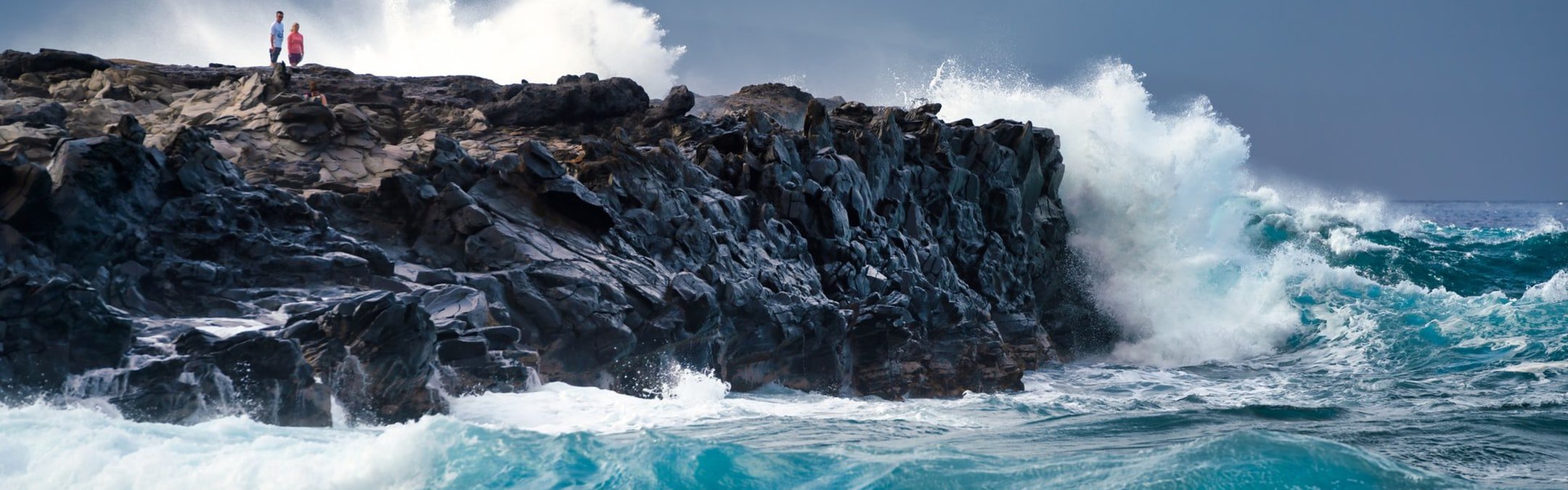 A shot of waves crashing into a cliff.On the cliff, 2 people can be seen.