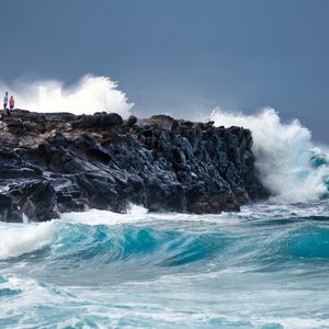 A shot of waves crashing into a cliff.On the cliff, 2 people can be seen.