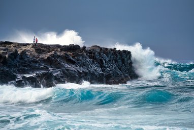 A shot of waves crashing into a cliff.On the cliff, 2 people can be seen.