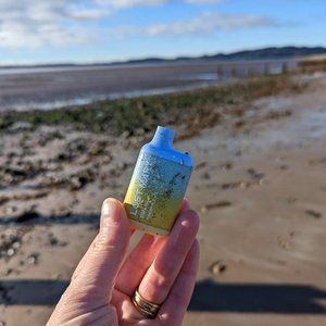 A hand is holding up a colourful disposable vape, the sandy beach is in the background. The disposable vape is litter.