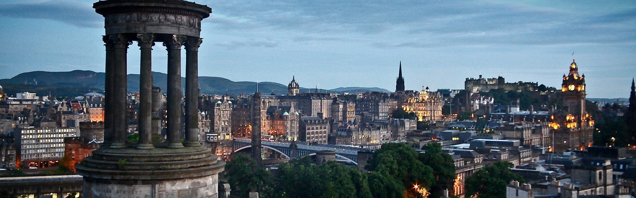 An evening view of the Edinburgh skyline from Calton Hill, featuring the Dugald Stewart Monument in the foreground and Edinburgh Castle illuminated in the distance under a twilight sky.
