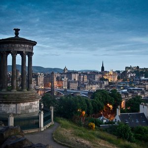 An evening view of the Edinburgh skyline from Calton Hill, featuring the Dugald Stewart Monument in the foreground and Edinburgh Castle illuminated in the distance under a twilight sky.