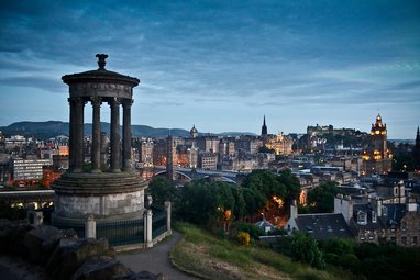 An evening view of the Edinburgh skyline from Calton Hill, featuring the Dugald Stewart Monument in the foreground and Edinburgh Castle illuminated in the distance under a twilight sky.