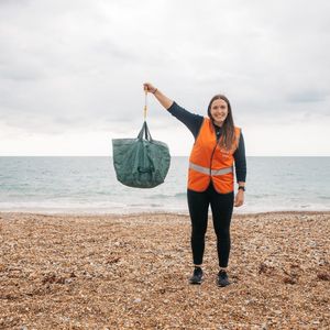 A woman in an orange hi-vis vest holding up a bag of litter with her right arm. She is standing on a pebble beach and is smiling.