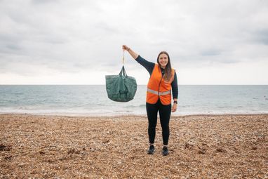 A woman in an orange hi-vis vest holding up a bag of litter with her right arm. She is standing on a pebble beach and is smiling.