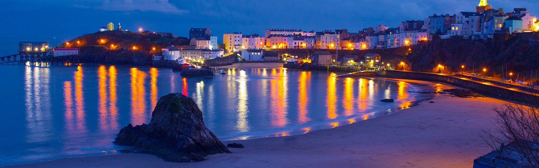 Tenby Harbour is captured at night time. The colourful lights from the buildings around the bay are reflected in the sea