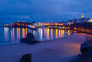 Tenby Harbour is captured at night time. The colourful lights from the buildings around the bay are reflected in the sea