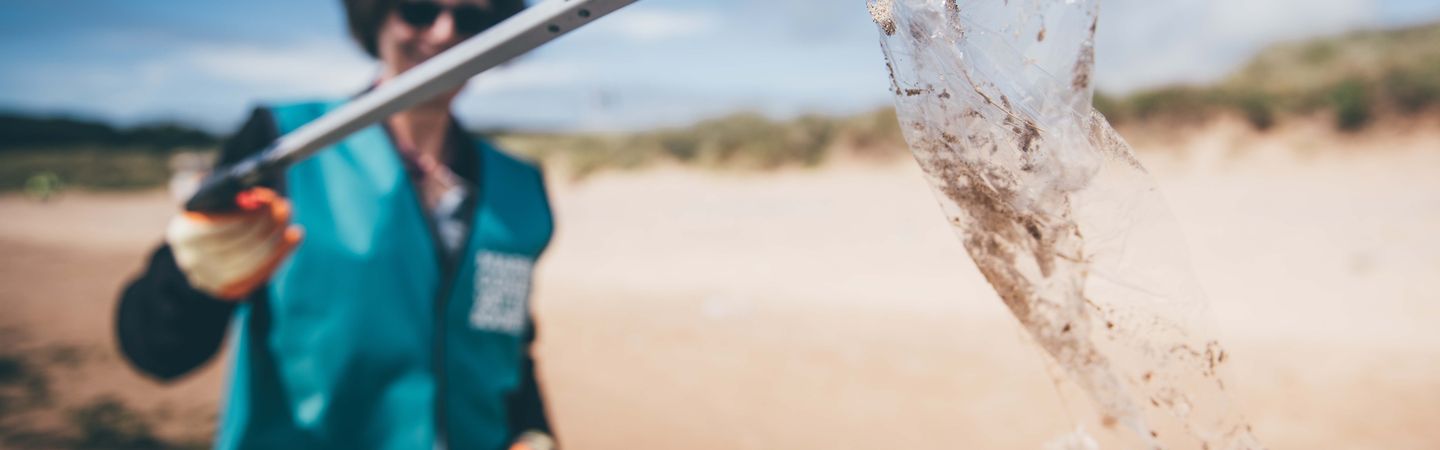 A woman holding up a piece of plastic wrapping using a litter picker to the camera. She is wearing a Marine Conservation Society vest. She is situated on a beach. The camera focus is on the wrapping making the woman blurry in the background.