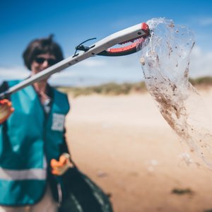 A woman holding up a piece of plastic wrapping using a litter picker to the camera. She is wearing a Marine Conservation Society vest. She is situated on a beach. The camera focus is on the wrapping making the woman blurry in the background.