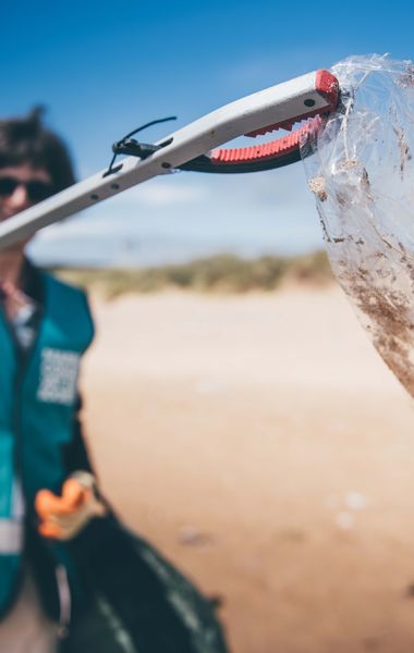 A woman holding up a piece of plastic wrapping using a litter picker to the camera. She is wearing a Marine Conservation Society vest. She is situated on a beach. The camera focus is on the wrapping making the woman blurry in the background.