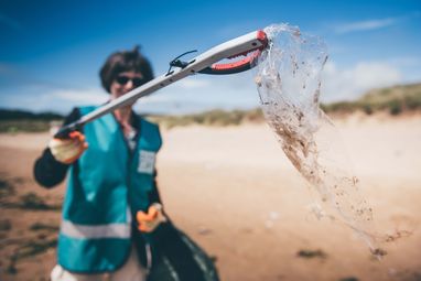 A woman holding up a piece of plastic wrapping using a litter picker to the camera. She is wearing a Marine Conservation Society vest. She is situated on a beach. The camera focus is on the wrapping making the woman blurry in the background.