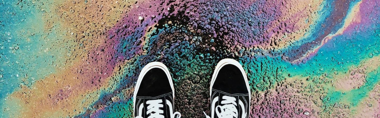The camera is looking down at a pair of shoes on the tarmac surrounded by a rainbow effect from oil on the road