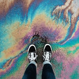 The camera is looking down at a pair of shoes on the tarmac surrounded by a rainbow effect from oil on the road