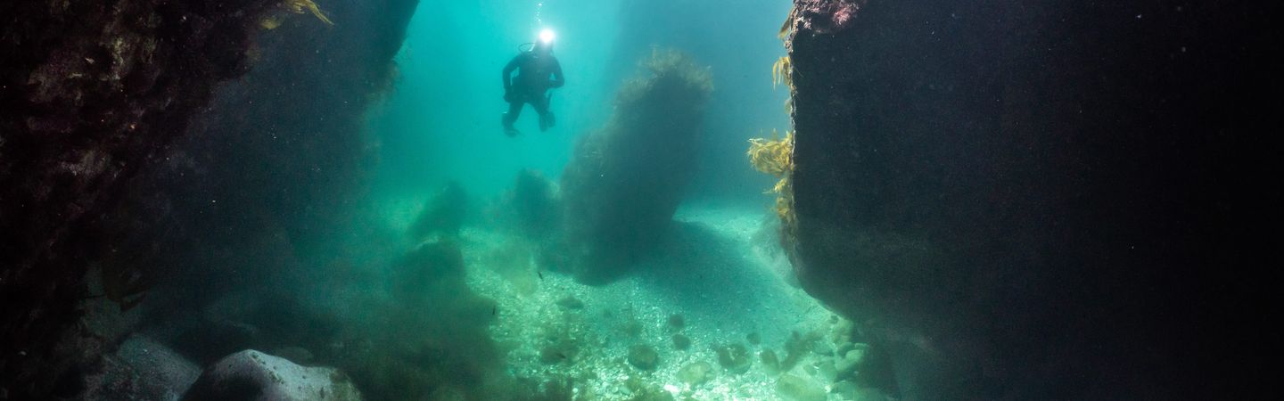 A scuba diver lighting up a dark underwater cave with a head torch, as sunlight filters down from the surface
