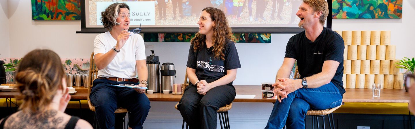 Zoe Lyons, a Marine Conservation Society staff member, and Cully from Cully and Sully, sit on stools in front of a large screen, giving a talk to an audience. All three people are smiling broadly.