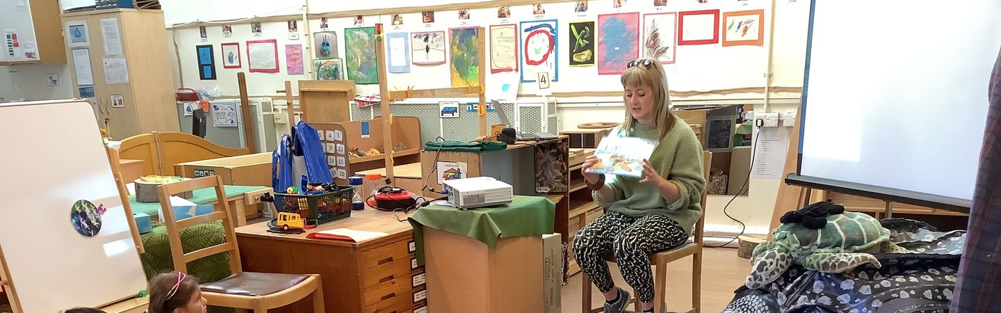 A member of Marine Conservation Society reading a book about a sea turtle to a group of young children sitting on the floor in a classroom.