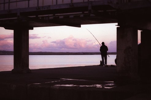 A silhouette of a lone fisherman under a pier.