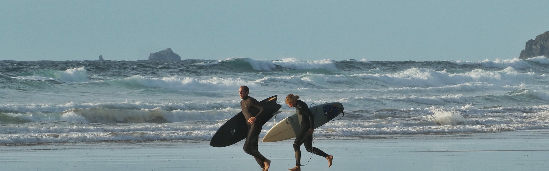 2 surfers holding surfboards and wearing wetsuits running alonga beach. The ocean is behind them