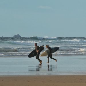 2 surfers holding surfboards and wearing wetsuits running alonga beach. The ocean is behind them