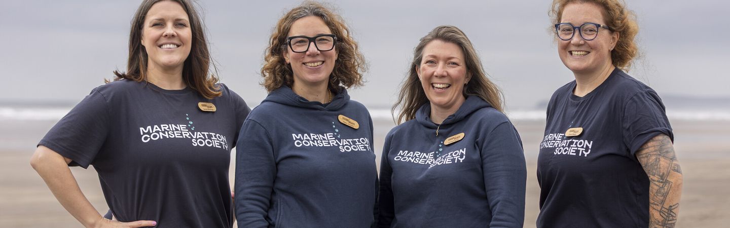 Four Marine Conservation Society staff stand together on a beach, smiling facing the camera, as they get ready to take part in a beach clean
