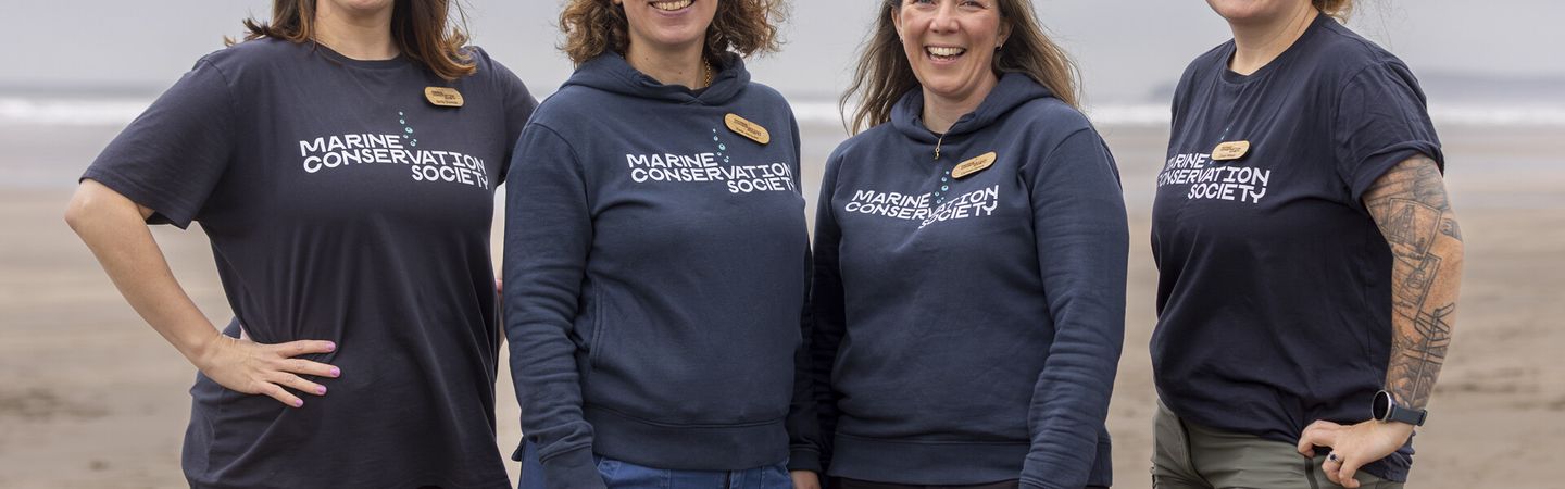 Four Marine Conservation Society staff stand together on a beach, smiling facing the camera, as they get ready to take part in a beach clean