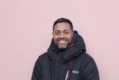 Trustee Rajah smiles widely against a pale pink background. He is wearing a thick black jacket suitable for very cold conditions.