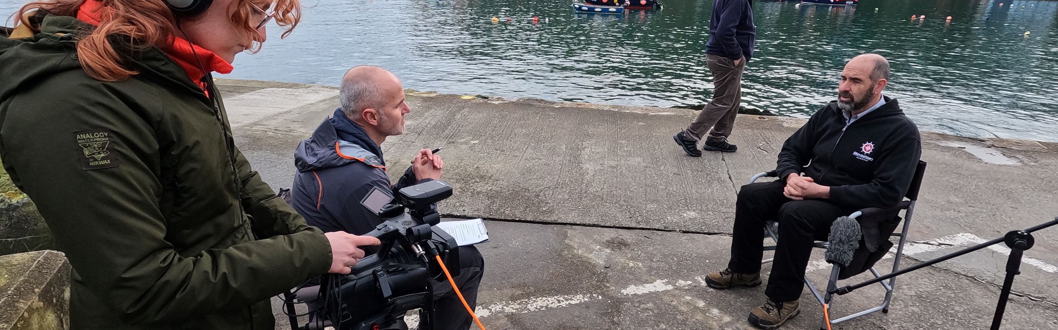 A film crew recording an interview with a man by the water in a busy harbor setting for the Valued Seas project