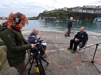 A film crew recording an interview with a man by the water in a busy harbor setting for the Valued Seas project