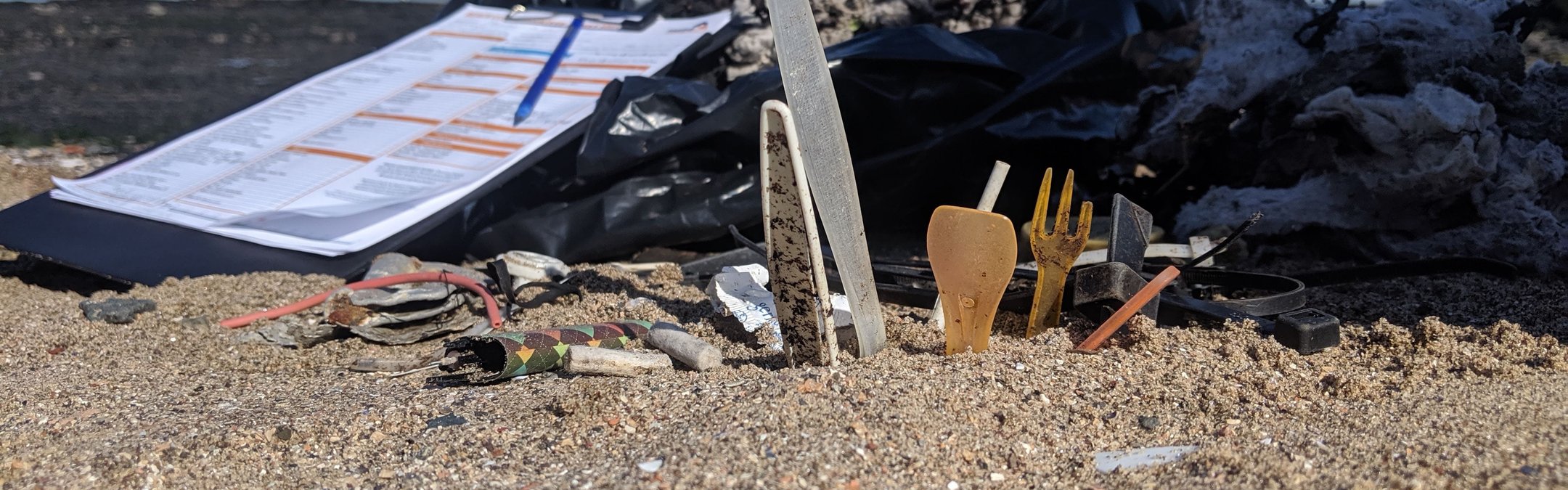 Single-use plastic on some sand. Next to it is a Marine Conservation Society beach clean survey form. The Forth bridge in Scotland can be seen behind it.
