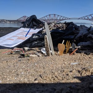 Single-use plastic on some sand. Next to it is a Marine Conservation Society beach clean survey form. The Forth bridge in Scotland can be seen behind it.