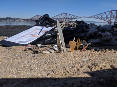 Single-use plastic on some sand. Next to it is a Marine Conservation Society beach clean survey form. The Forth bridge in Scotland can be seen behind it.