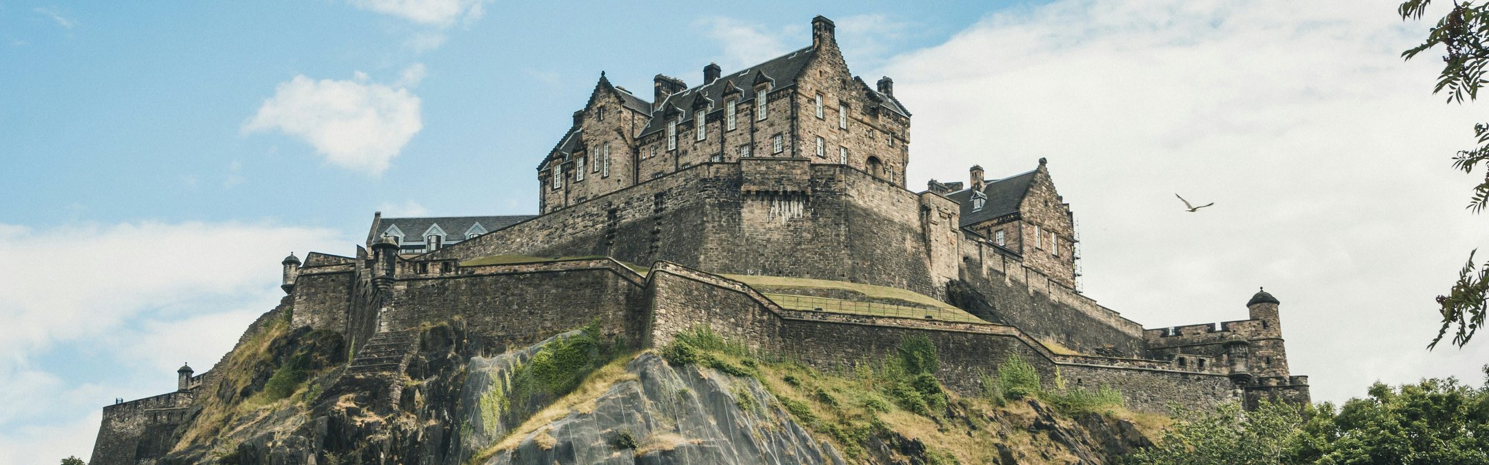 A low-angle shot of the historic Edinburgh Castle perched atop a jagged, rocky cliff in Scotland. The fortress stands prominently against a bright blue sky with wispy white clouds. Foliage frame the bottom of the image.