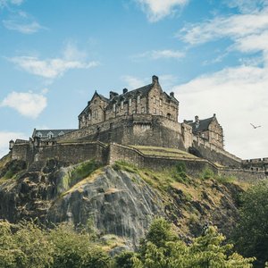 A low-angle shot of the historic Edinburgh Castle perched atop a jagged, rocky cliff in Scotland. The fortress stands prominently against a bright blue sky with wispy white clouds. Foliage frame the bottom of the image.