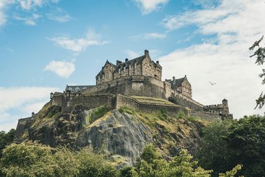 A low-angle shot of the historic Edinburgh Castle perched atop a jagged, rocky cliff in Scotland. The fortress stands prominently against a bright blue sky with wispy white clouds. Foliage frame the bottom of the image.