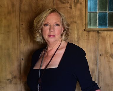 Deborah Meaden smiles at the camera in what looks like the industrial backdrop of the Dragon's Den studio. She is wearing a smart black dress.